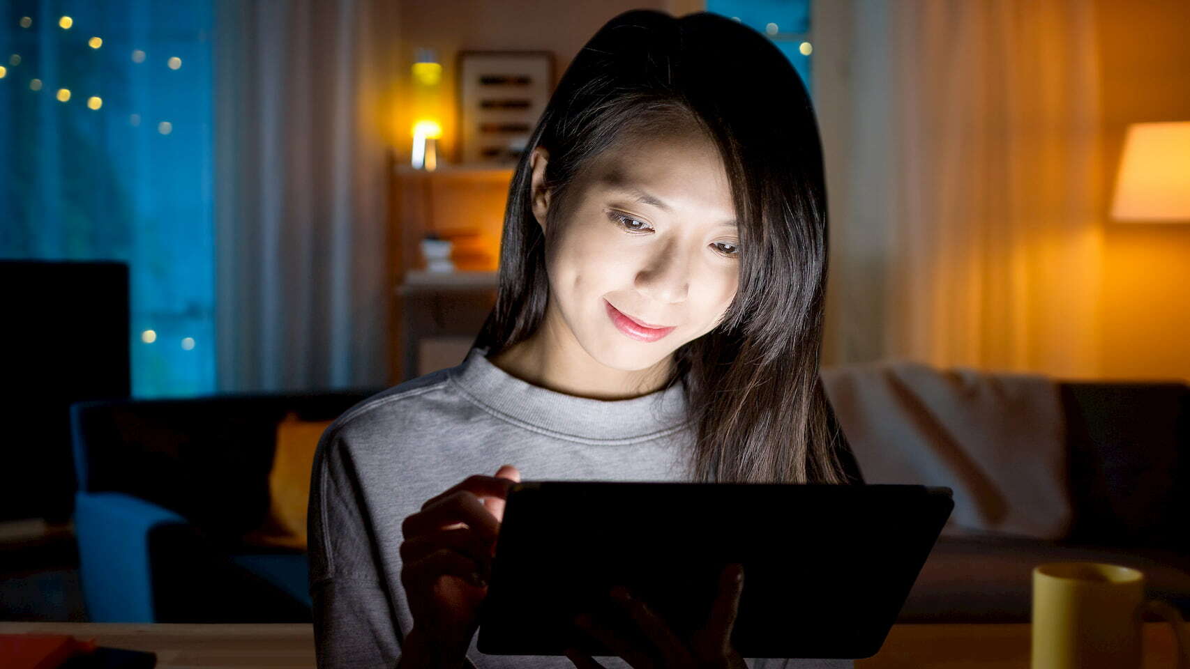 Photograph of a young Asian woman enjoying her tablet using wifi internet in her warmly lit living room on a cozy evening at home